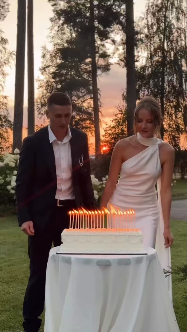 Couple standing behind a white rectangular cake with lit candles at sunset.