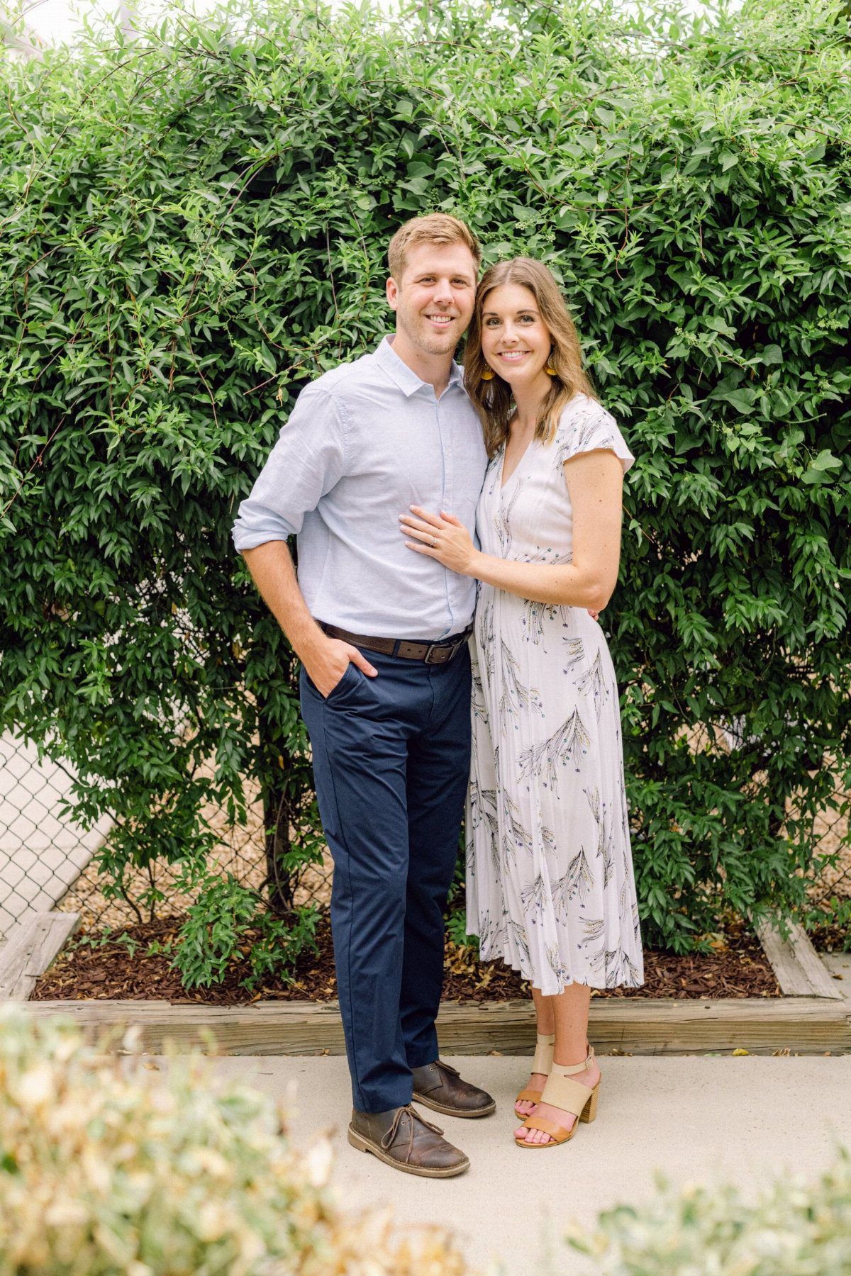 Man in blue shirt and woman in floral midi dress posing by green hedge.