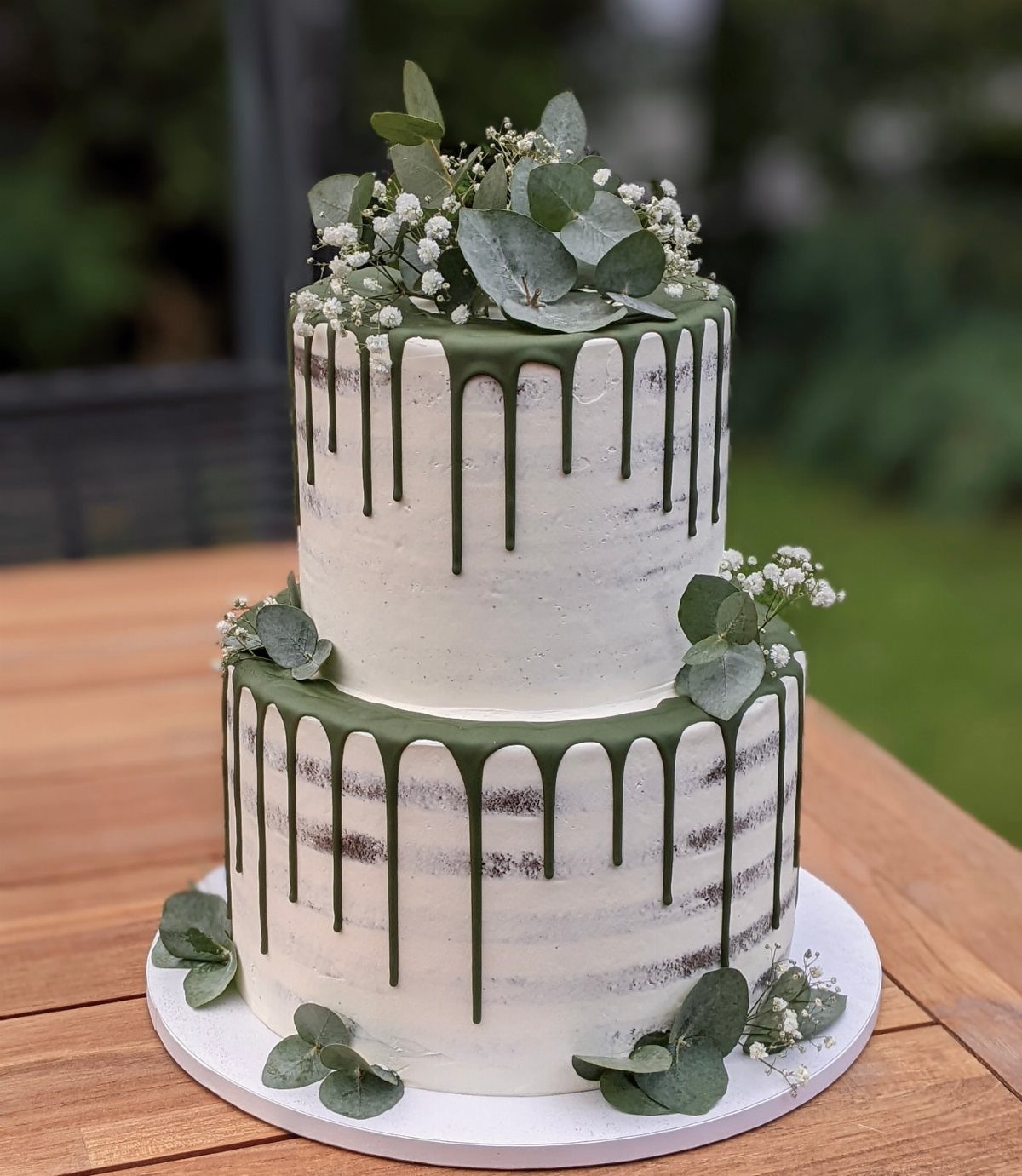 Two-tier semi-naked cake with green drip icing and eucalyptus greenery on wooden table.