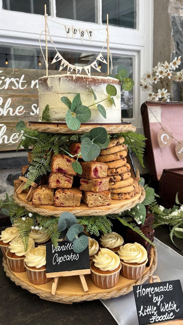 Semi-naked wedding cake with greenery on a tiered wooden stand with cupcakes.