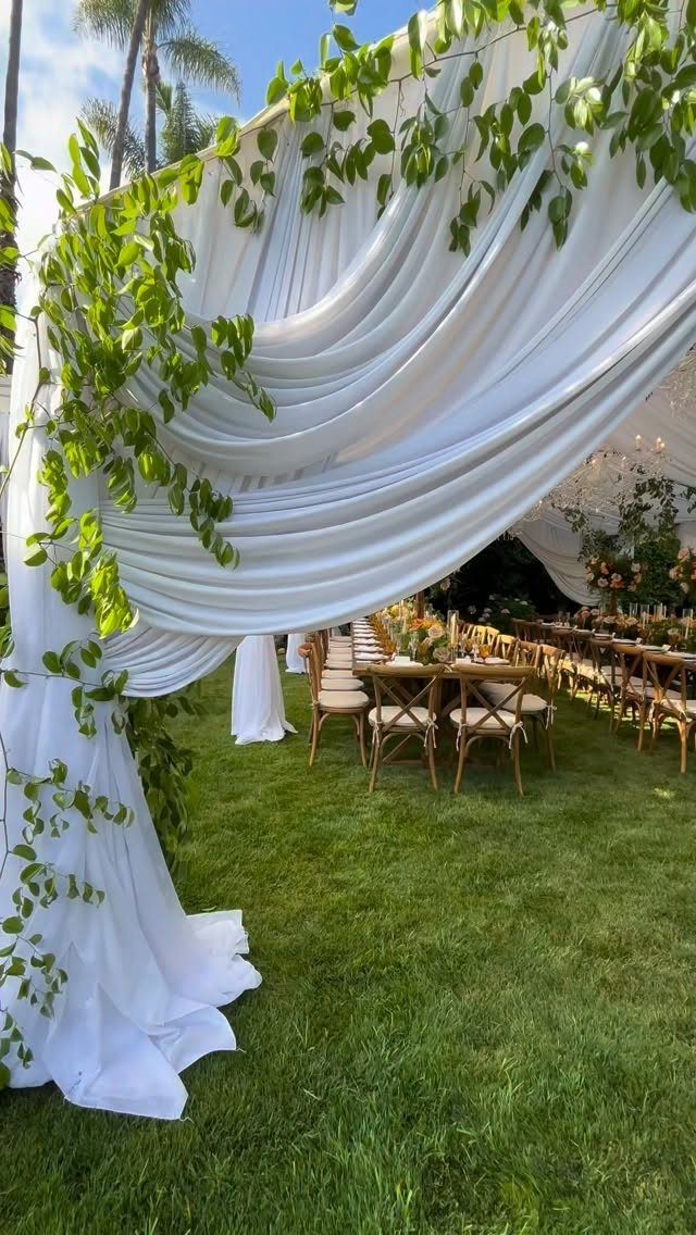 White draped fabric canopy with green vines over wooden tables on a grassy lawn.