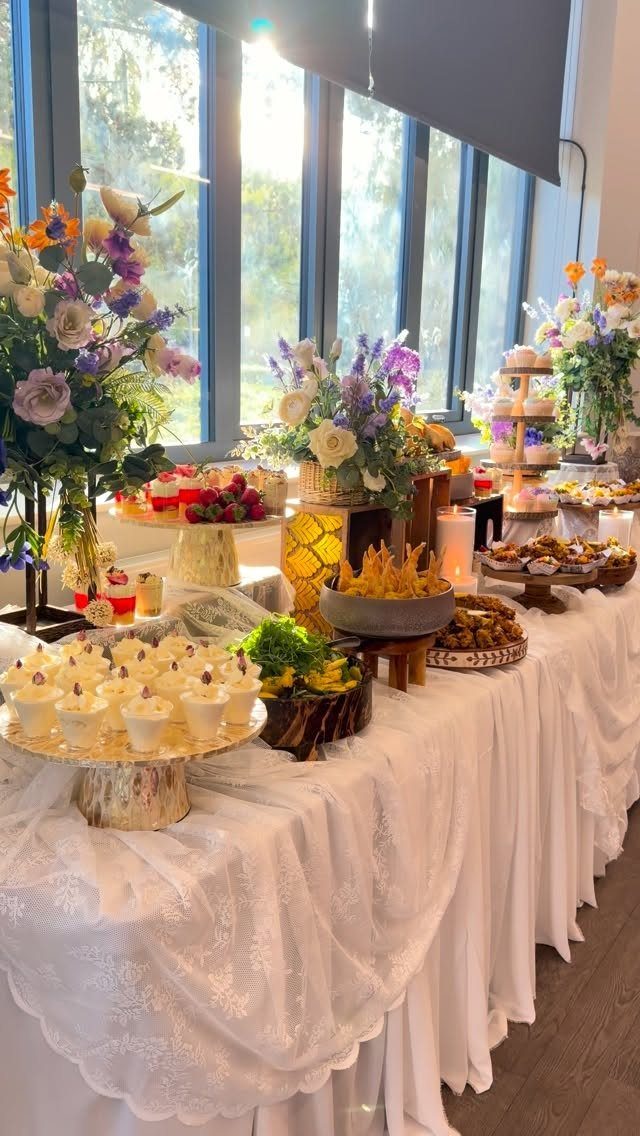 Buffet table with white lace cloth, cupcakes, appetizers, and colorful floral arrangements.