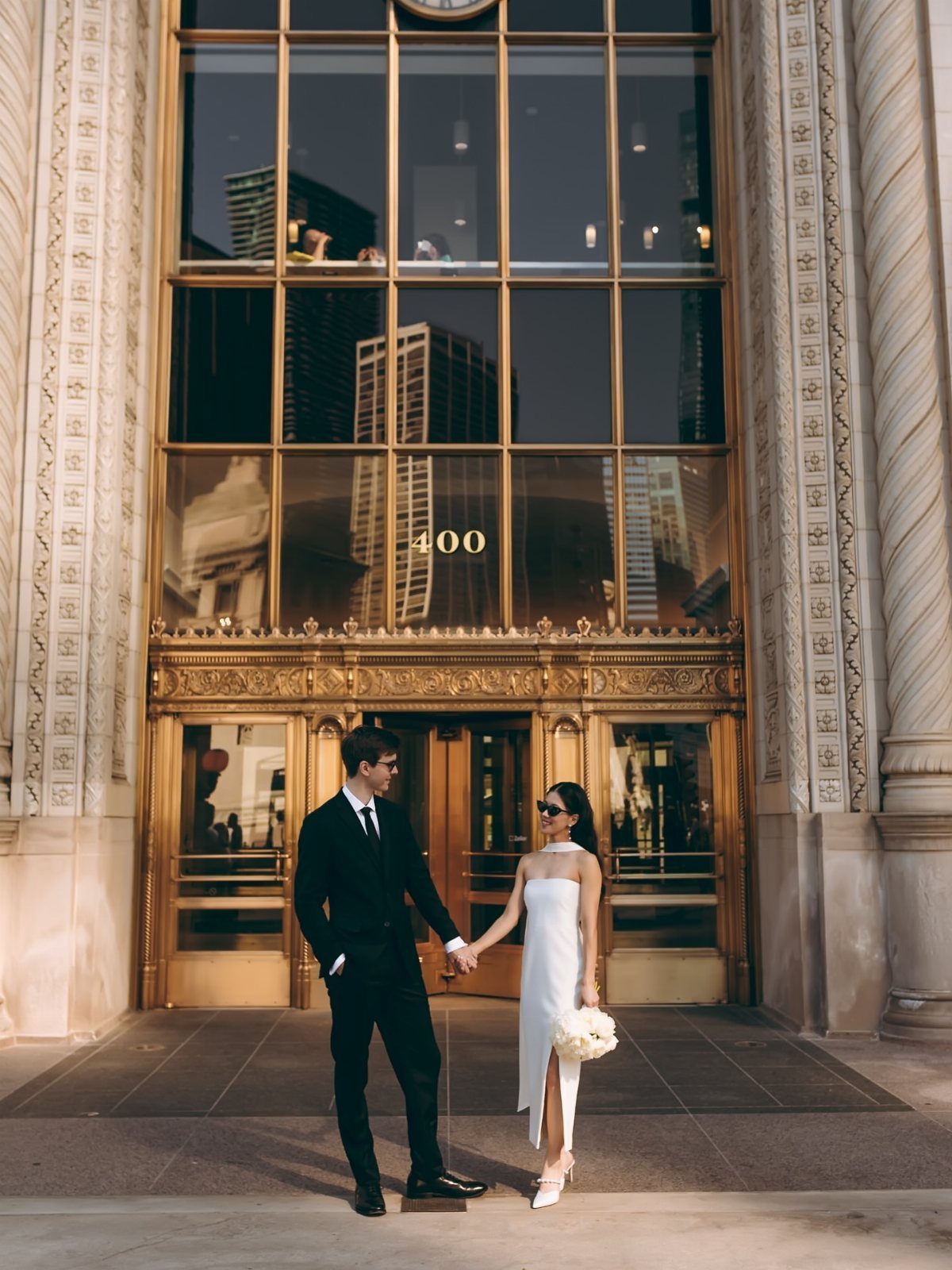 Couple in wedding attire holding hands before a grand city building entrance.