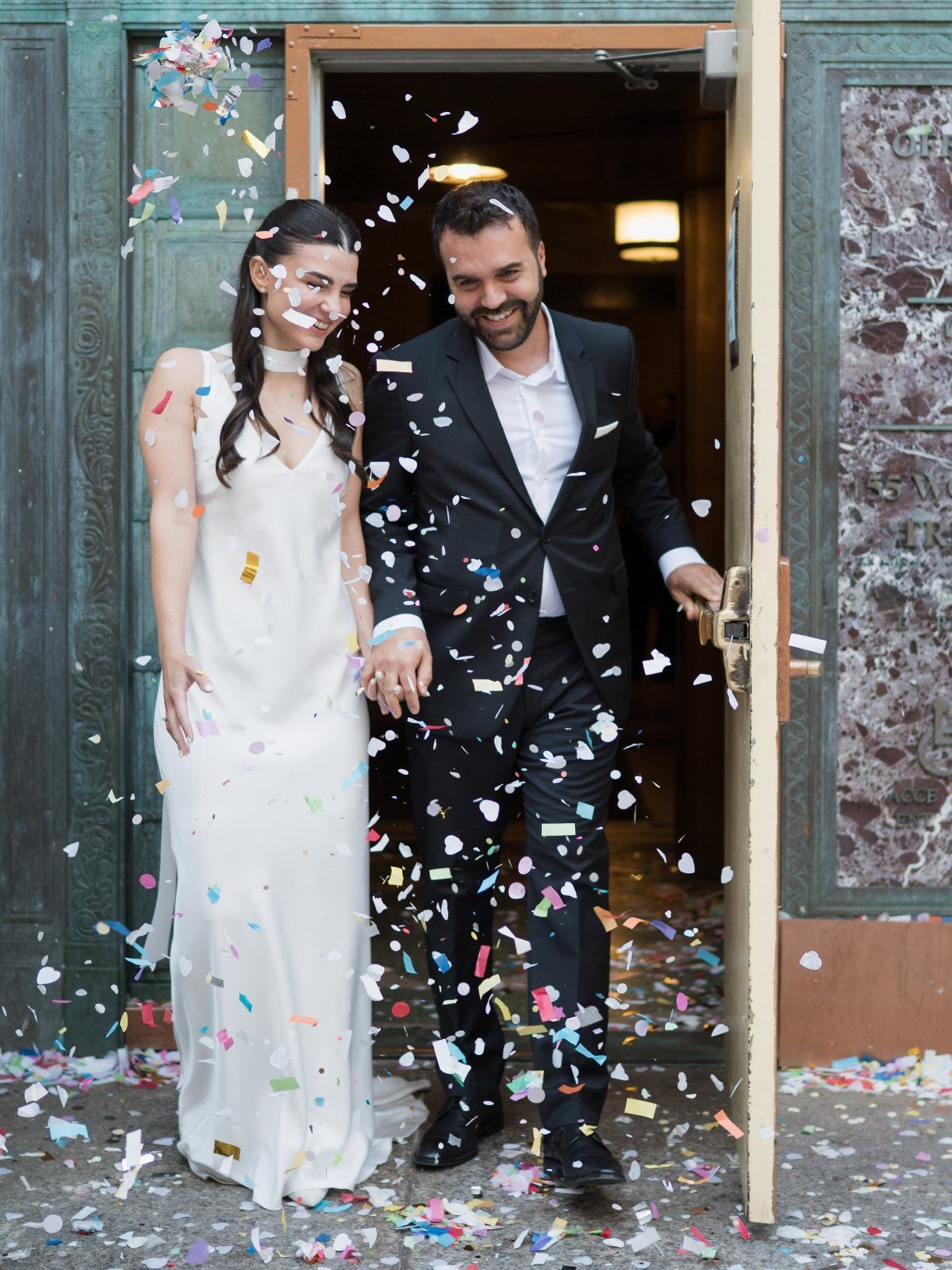 Newlywed couple smiling and walking through a doorway while colorful confetti falls around them.
