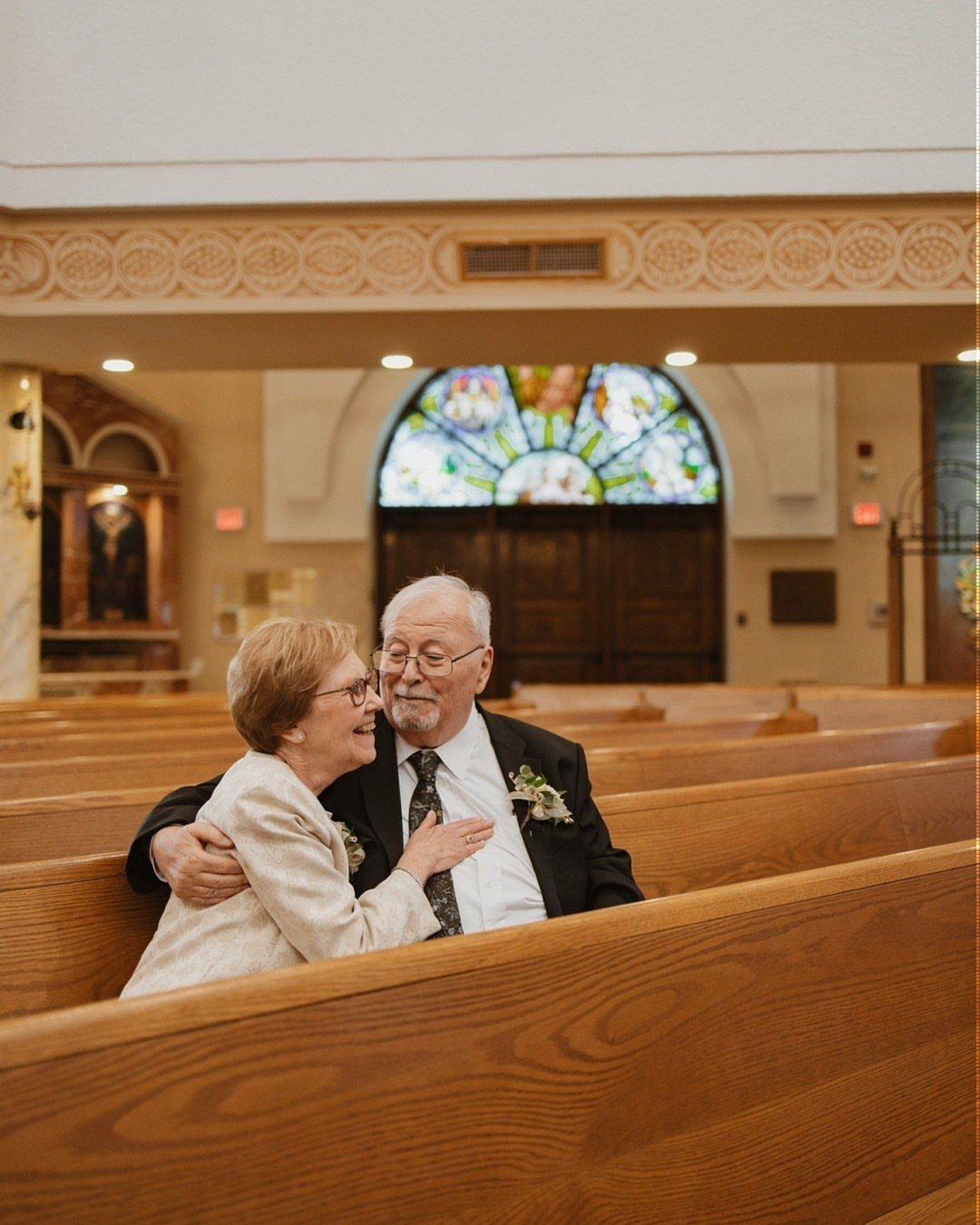 Elderly couple in formal wear embracing inside a church with wooden pews.