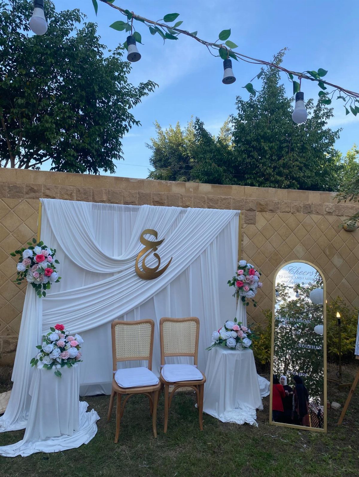 White draped wedding backdrop with gold ampersand, floral arrangements, and two wooden chairs.