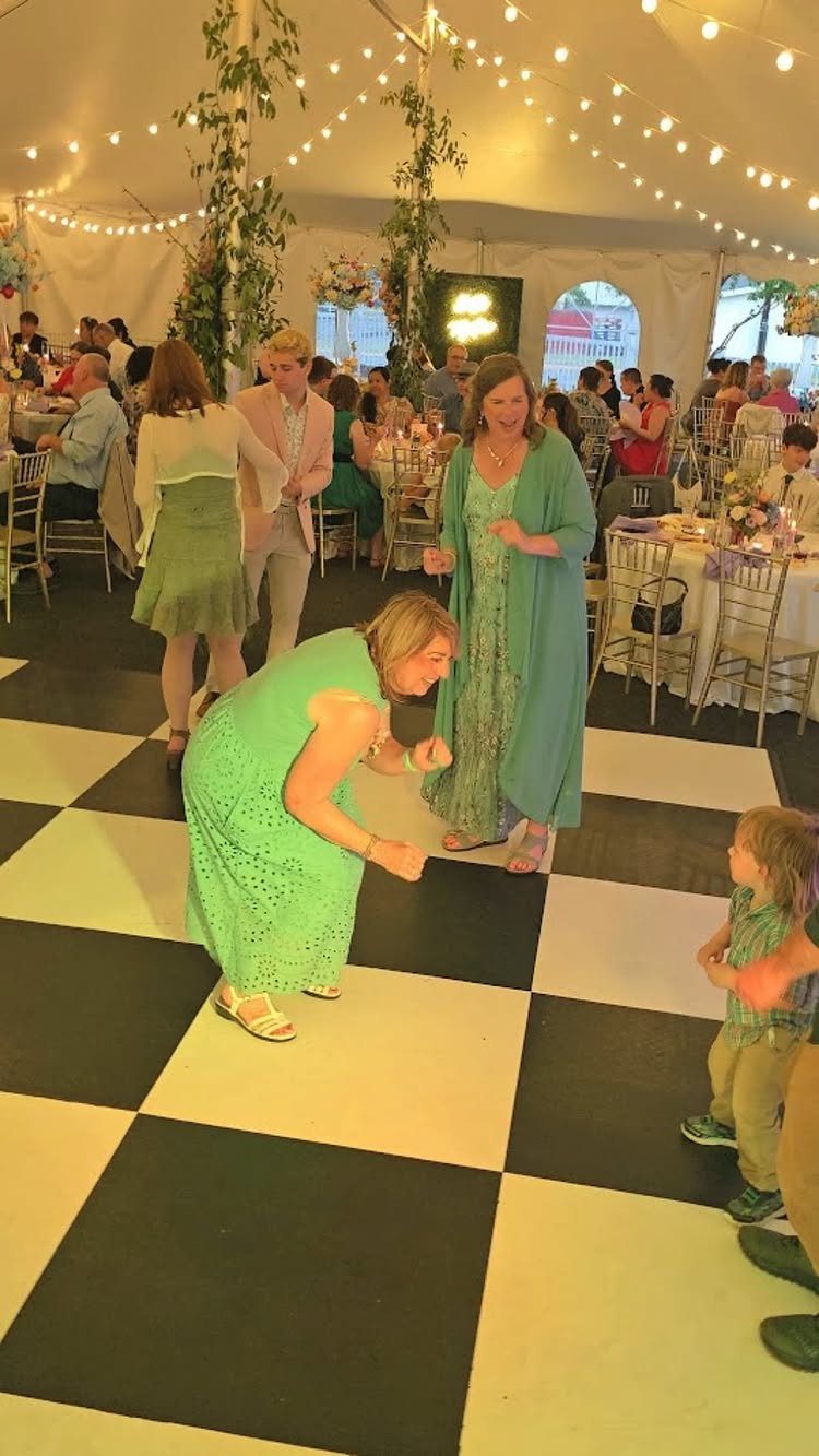 Guests dancing on a checkered floor at a wedding reception inside a lit tent.