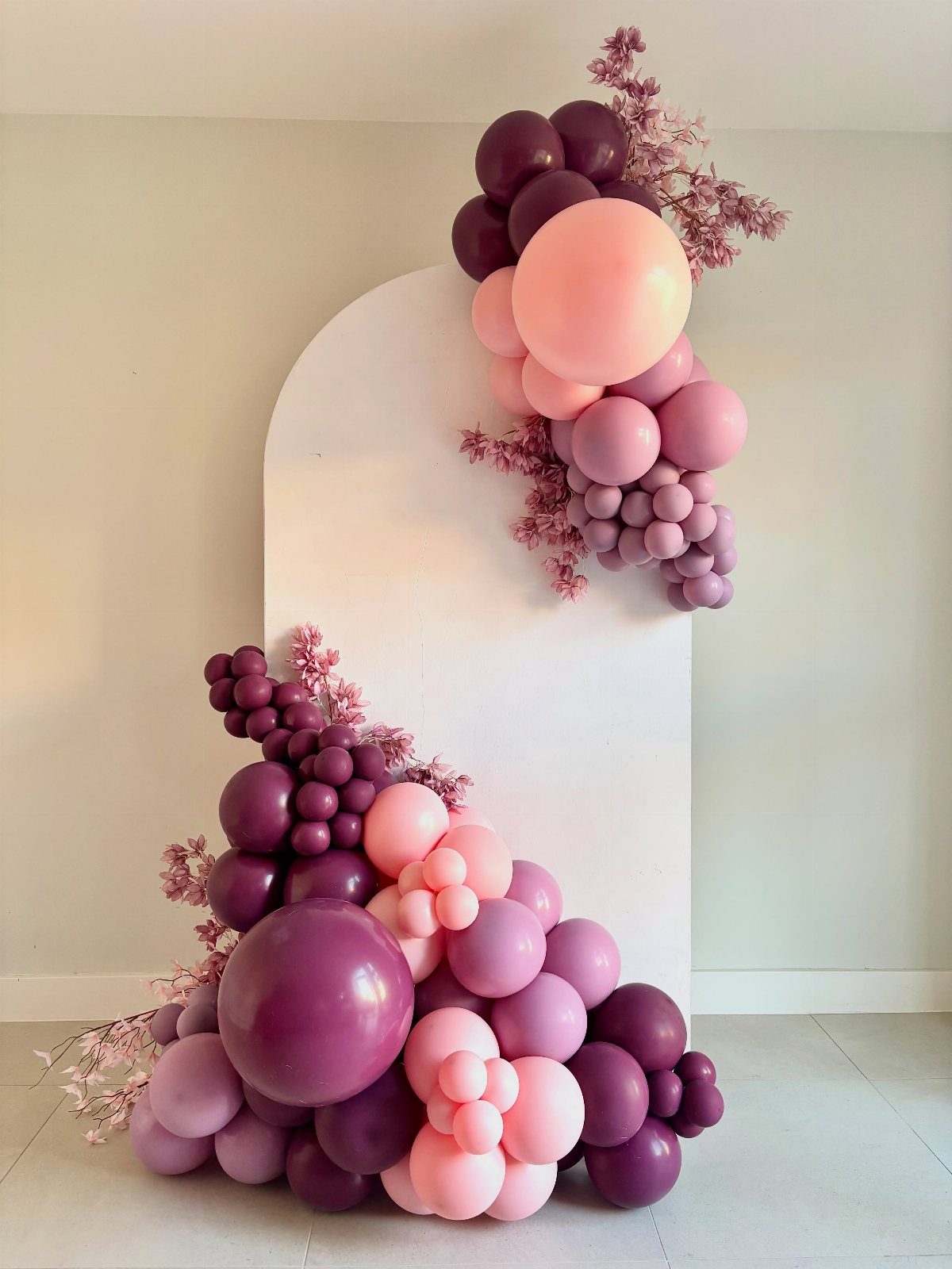 White arched backdrop decorated with purple and pink balloon garlands and floral accents.