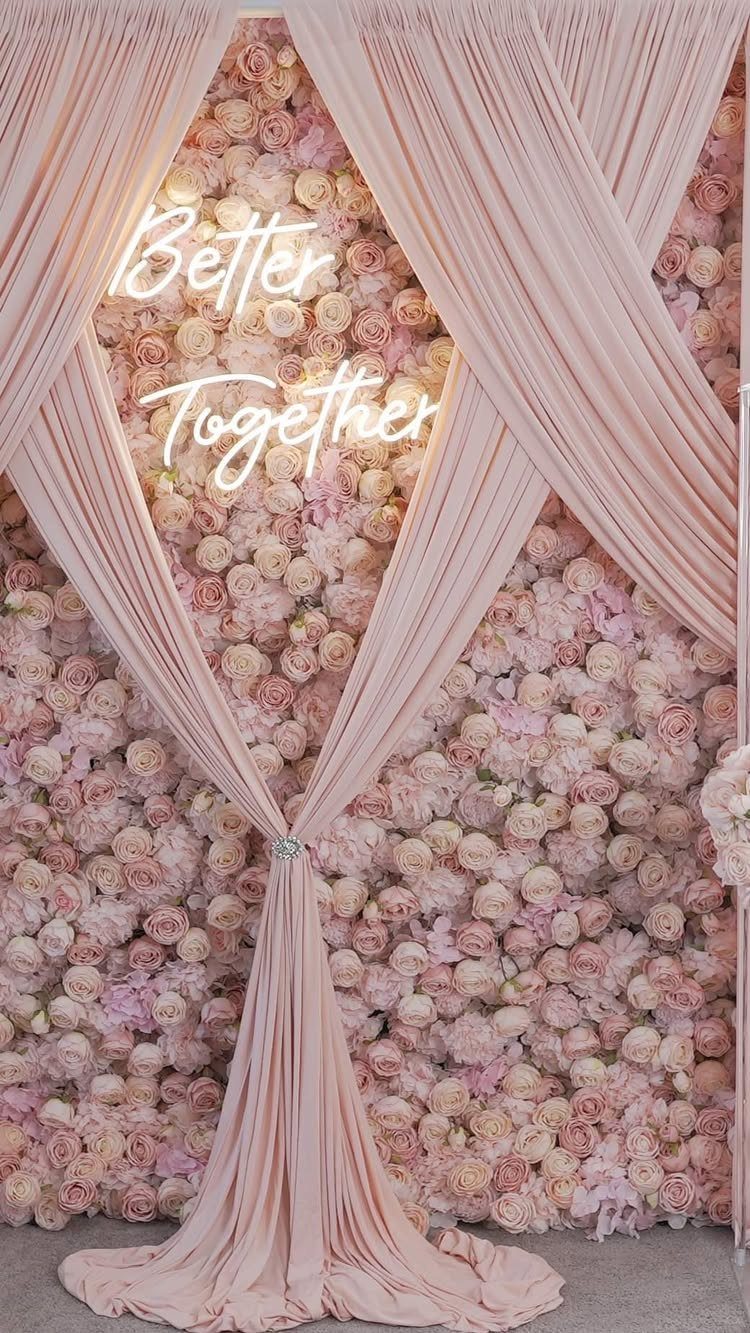 Artificial flower wall with pink roses, draped fabric, and a neon sign.