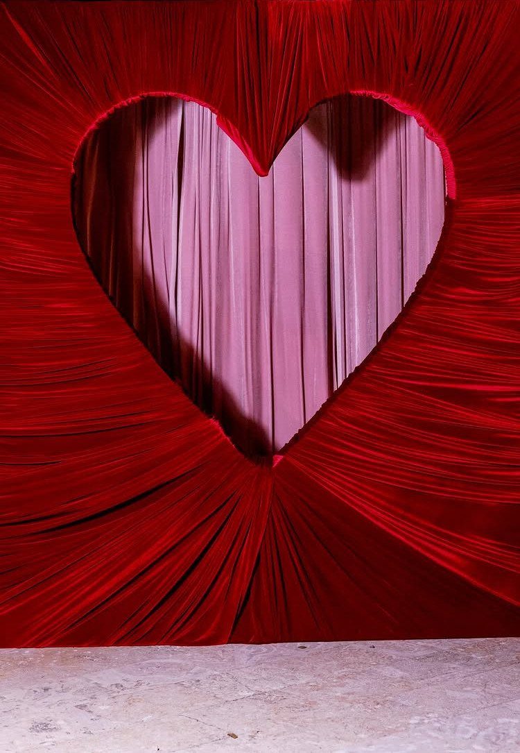 Large heart-shaped cutout in red velvet fabric with pink curtains behind it.