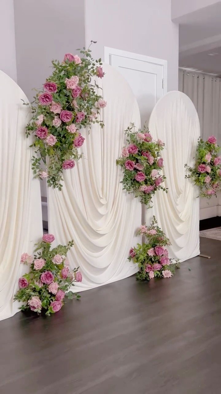 White draped fabric arched backdrops adorned with pink and purple floral arrangements.