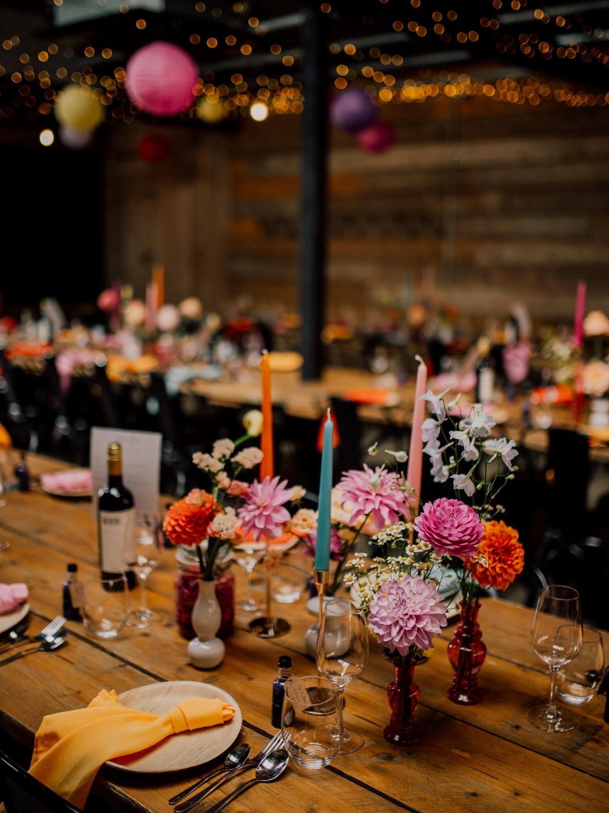 Wooden wedding table with colorful flower centerpieces, tapered candles, and overhead string lights.