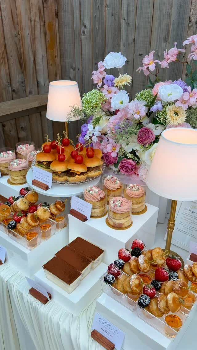 Dessert table with mini cakes, pastries, and sliders decorated with flowers and lamps.