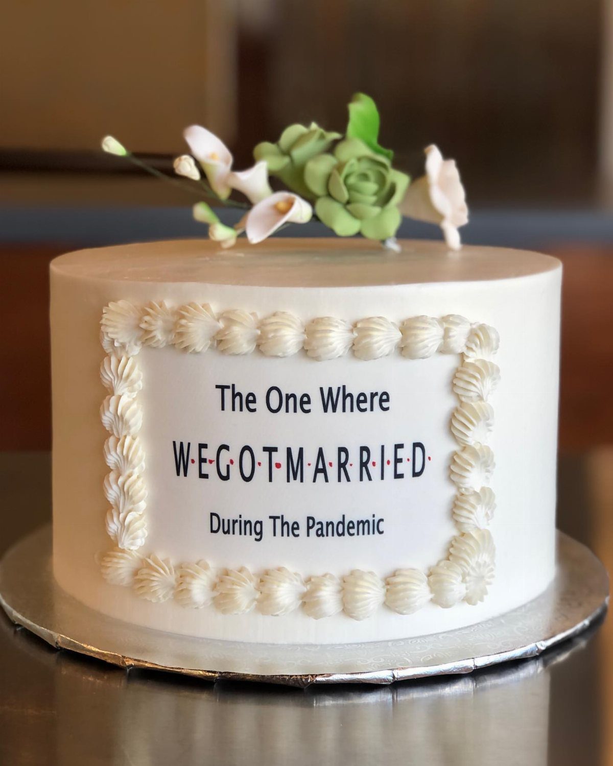 White wedding cake with a Friends-themed plaque and small sugar flower toppers.