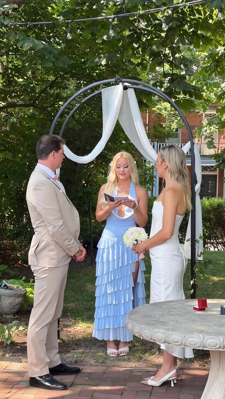 Woman in blue dress officiating a wedding ceremony under a white fabric arch.