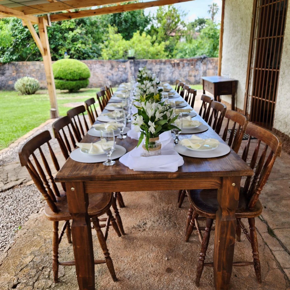 Long wooden dining table set with white plates and lilies in a garden.