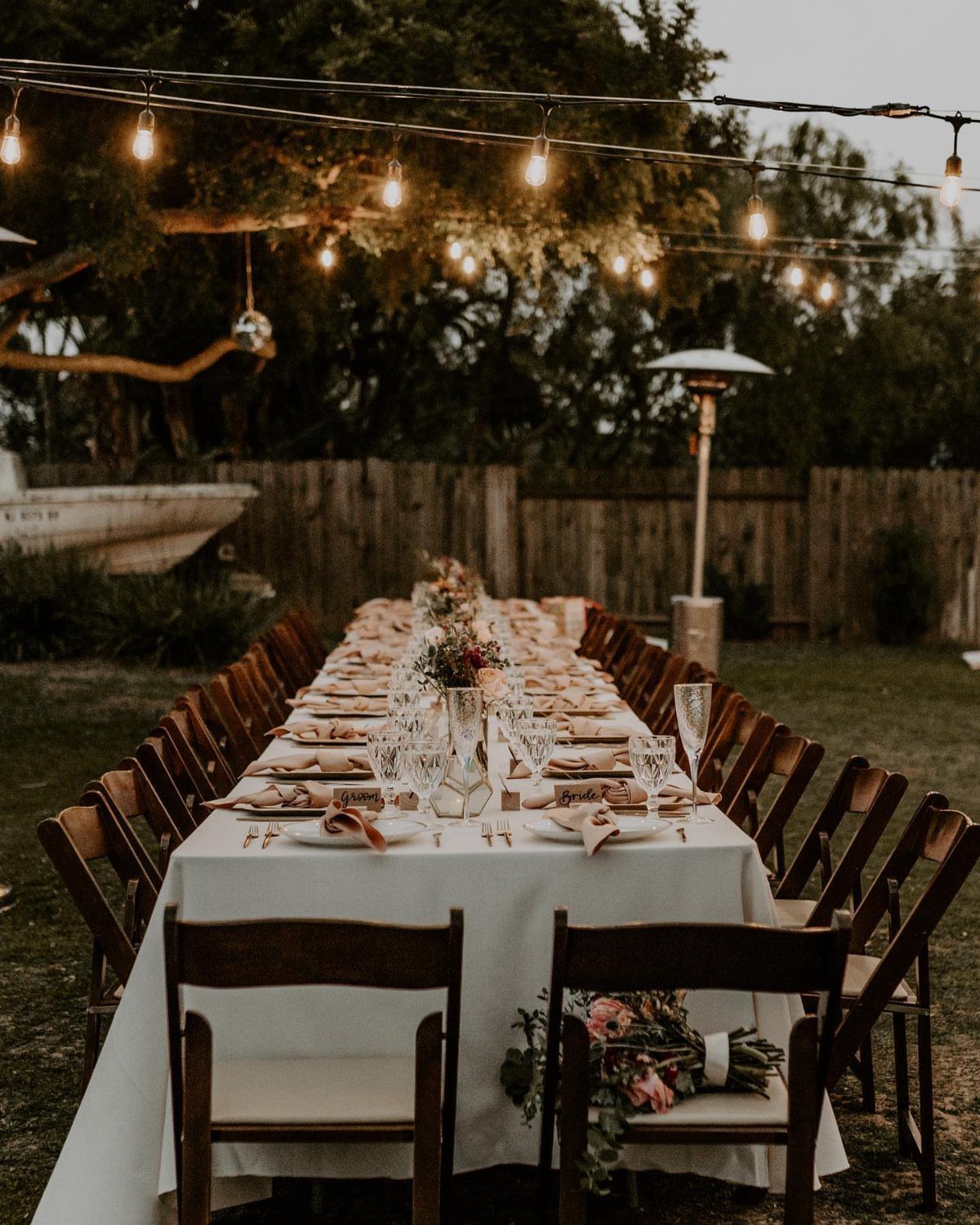 Long outdoor wedding table with white linens, floral centerpieces, and overhead string lights.