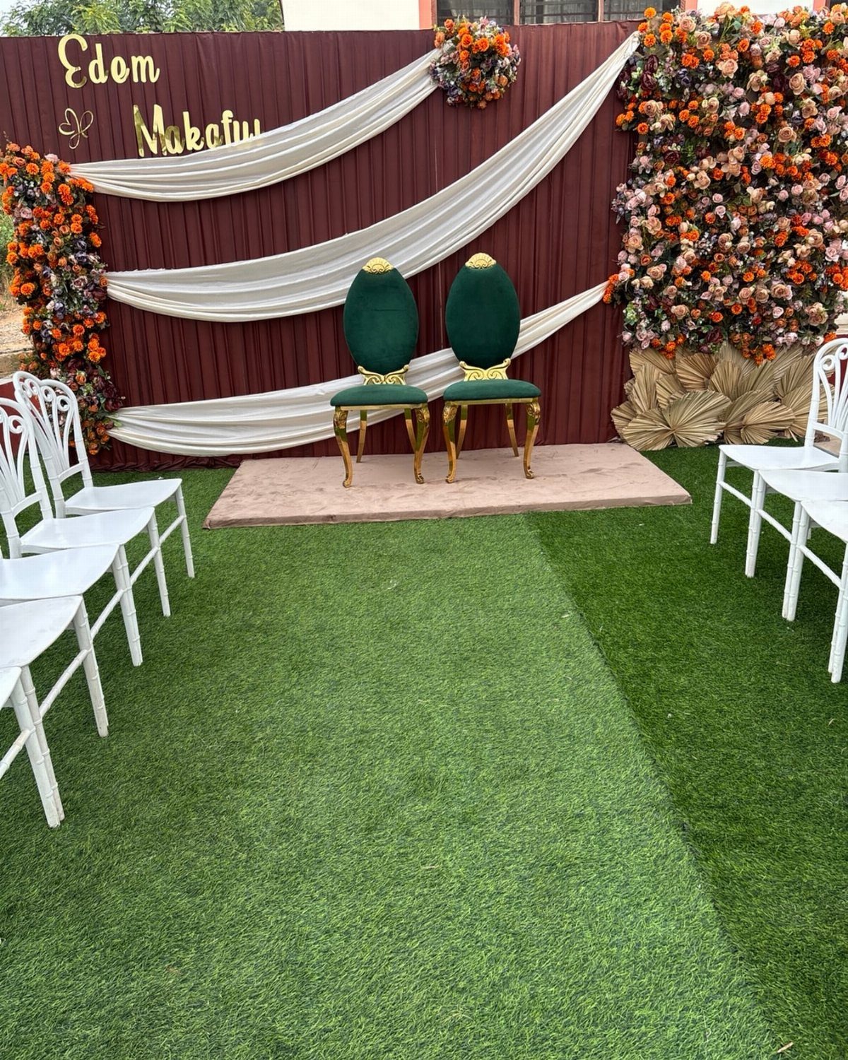 Ceremony backdrop with white fabric drapes, orange flowers, and green velvet chairs on grass.