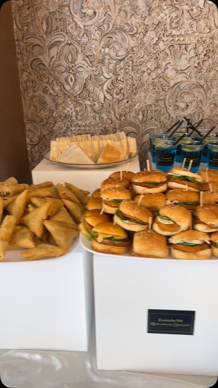 Platters of sliders and samosas on white pedestals against an ornate carved wall.