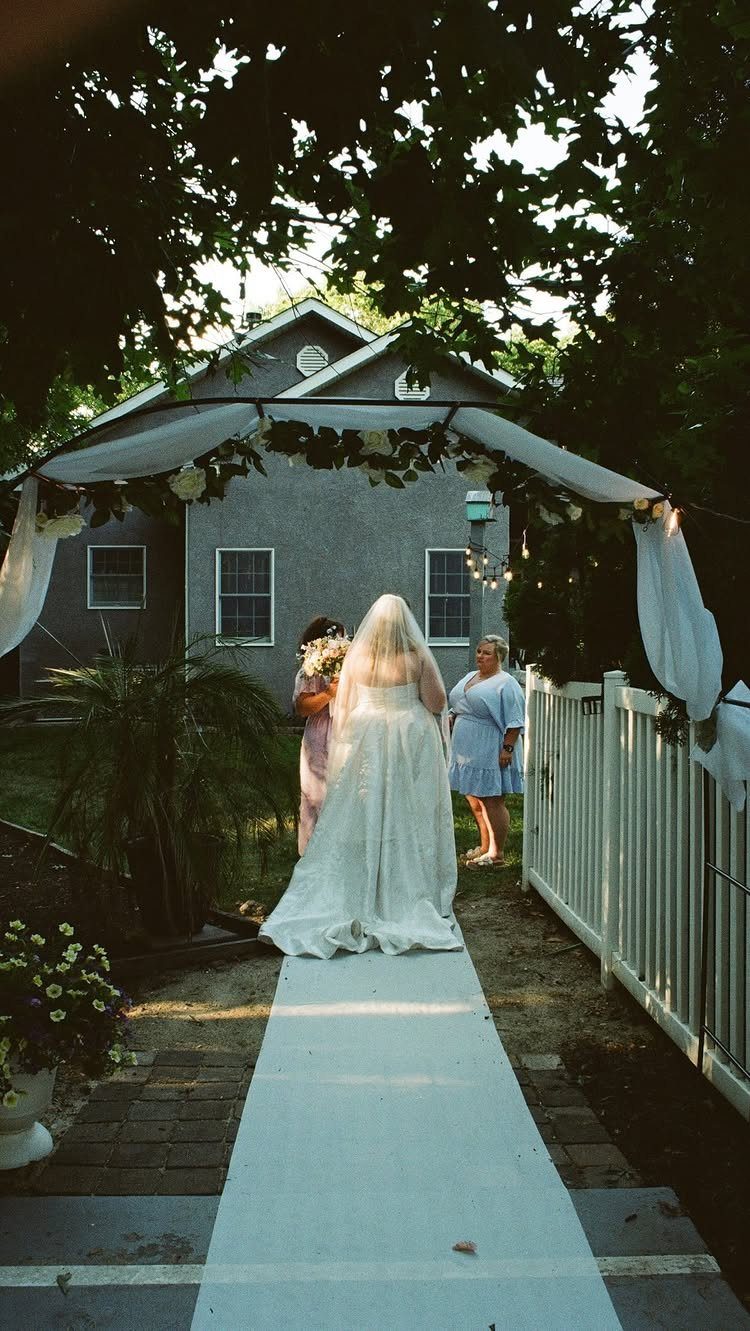 Bride in white gown walking down a white runner toward a backyard ceremony arch.