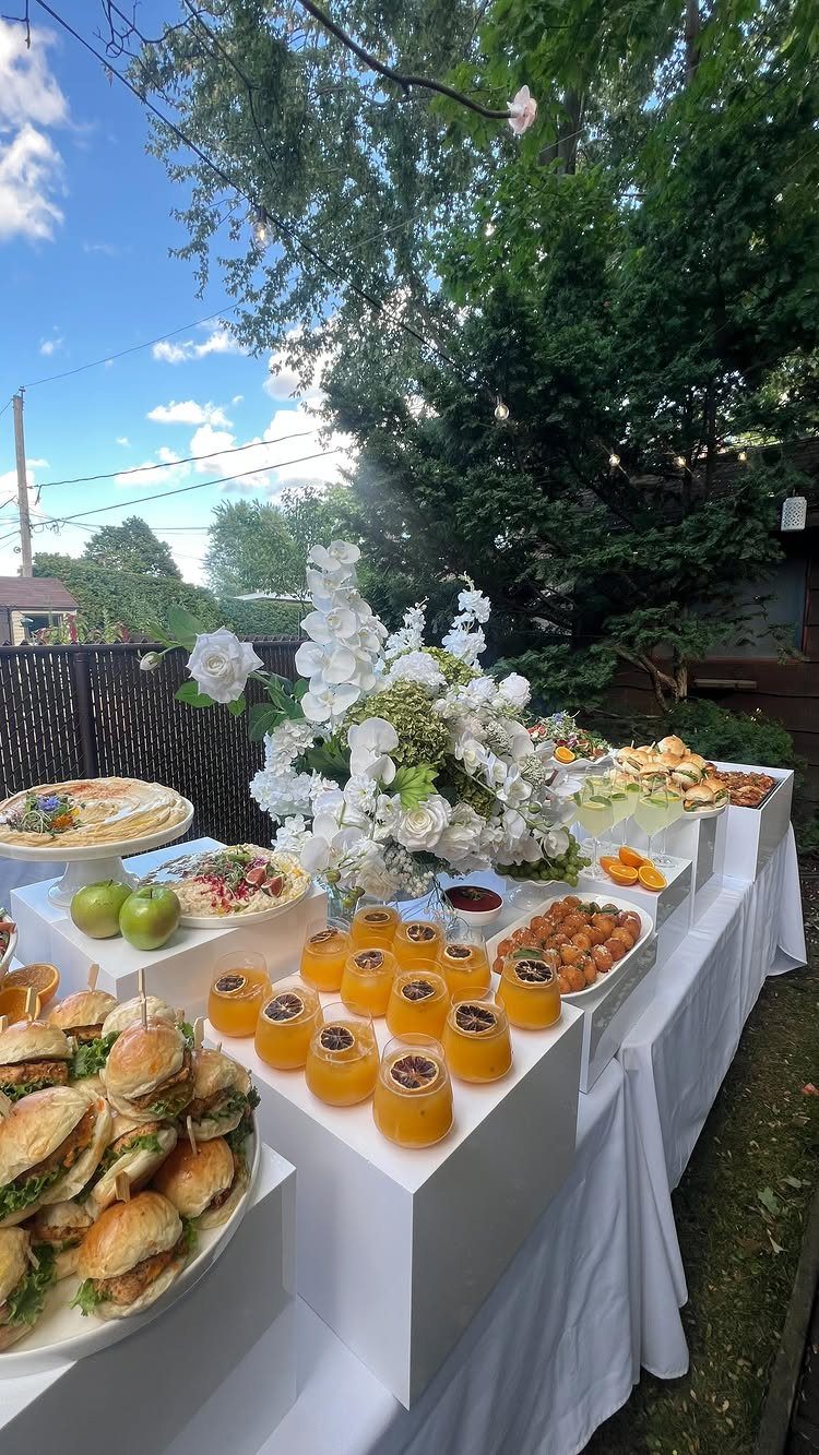 Brunch food spread with sliders, orange juice, and white flowers on white tables outdoors.
