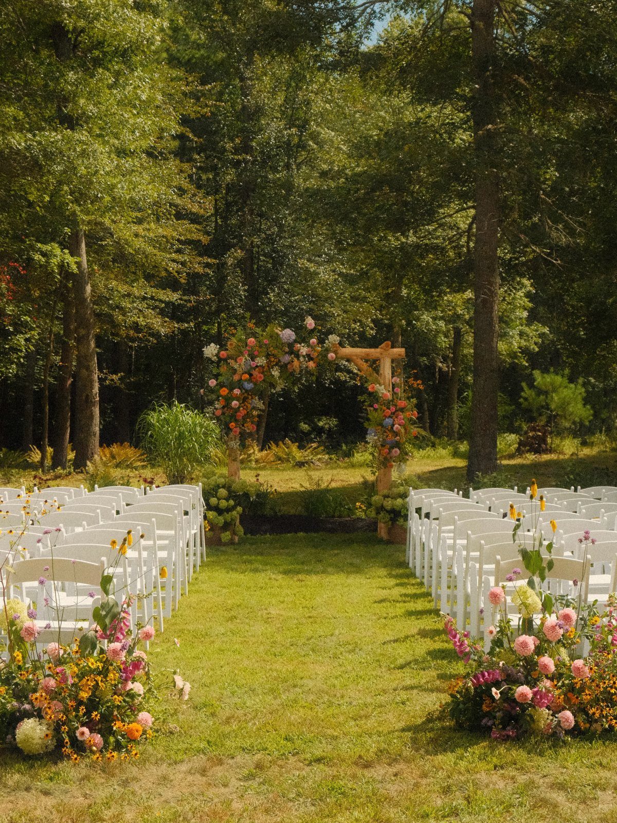Outdoor wedding ceremony setup with white chairs and a floral wooden arch in a forest.