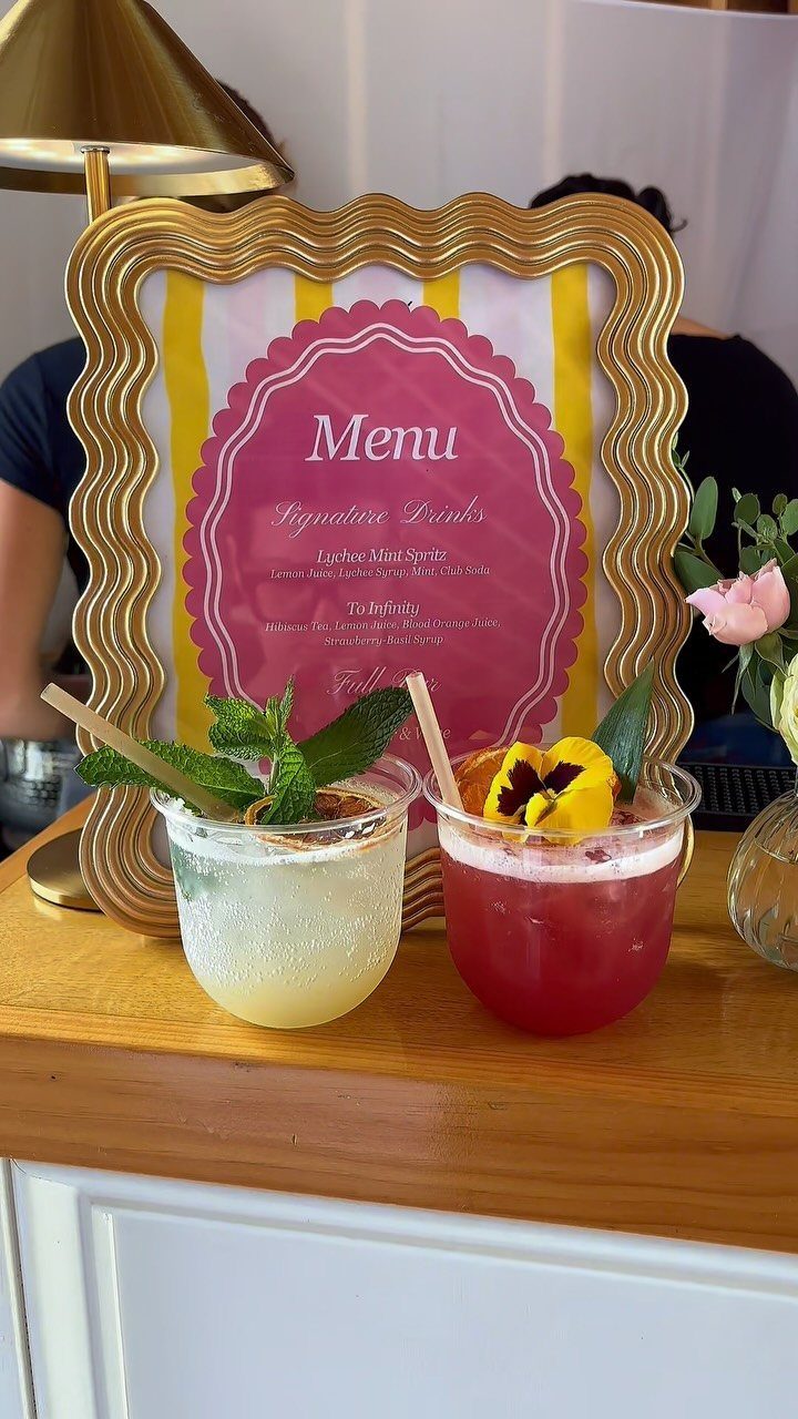 15 Small Wedding Ideas for 50 Guests, From Backyards to Parks 10 Two garnished cocktails on a wooden counter next to a gold-framed menu.