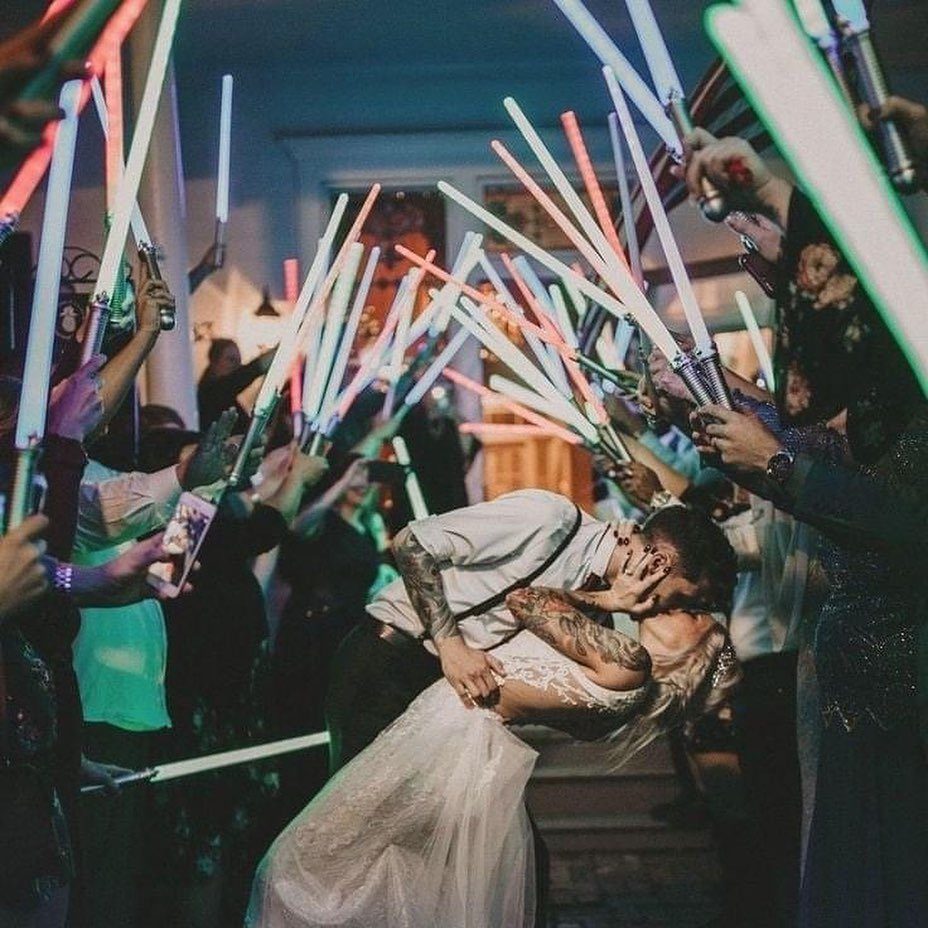 Bride and groom kissing under an arch of glowing colorful lightsabers held by guests.