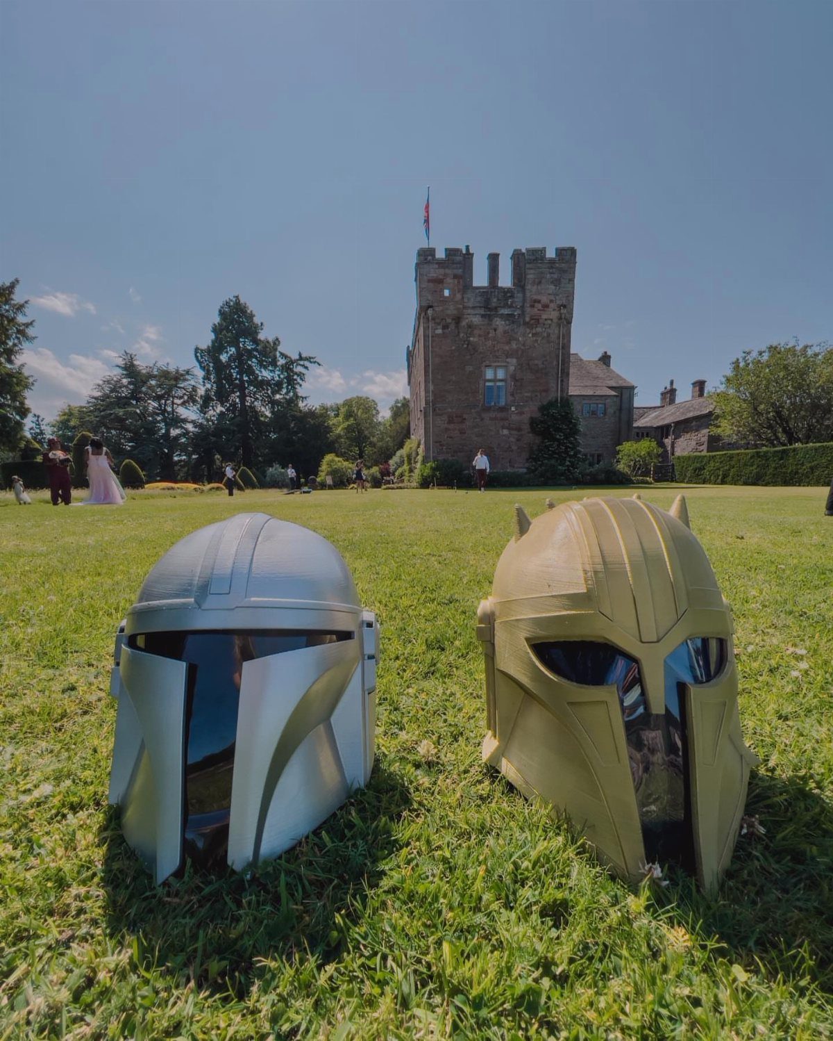 Two Mandalorian helmets on grass with a castle and bride in the background.