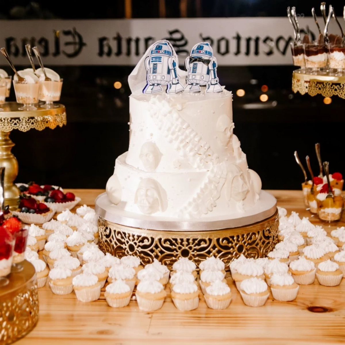 White wedding cake with R2-D2 toppers surrounded by white cupcakes and fruit tarts.