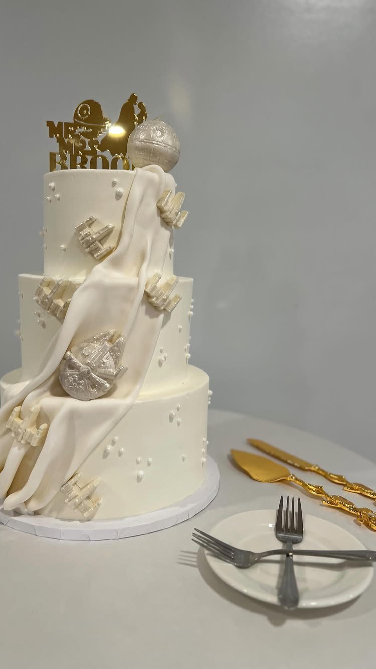 Three-tier white wedding cake with Death Star and Stormtrooper decorations and gold topper.