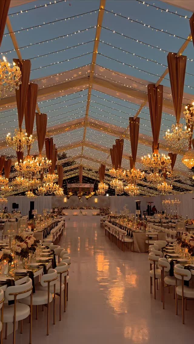 16 Tent Wedding Ideas for Lighting, Tables and Cozy Corners 12 Ornate gold chandeliers hanging inside a clear-top wedding tent over elegant dining tables.