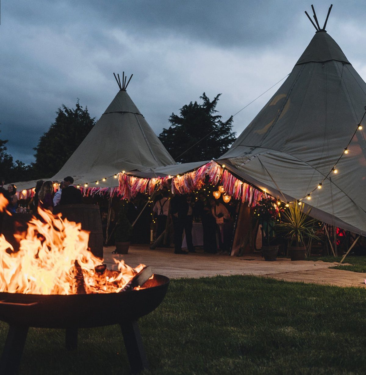 16 Tent Wedding Ideas for Lighting, Tables and Cozy Corners 6 Burning fire pit in foreground with decorated tipis and guests in background
