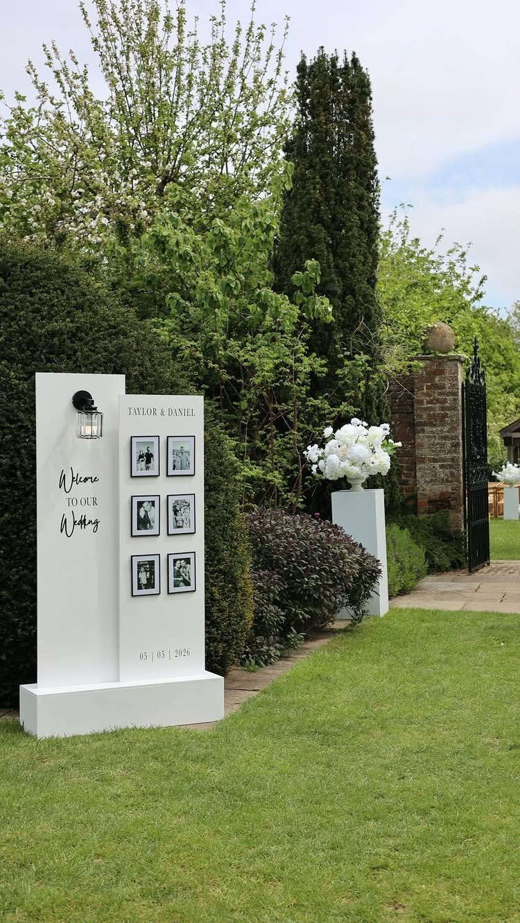 16 Tent Wedding Ideas for Lighting, Tables and Cozy Corners 3 White wedding welcome sign with photos standing on a green lawn by hedges.
