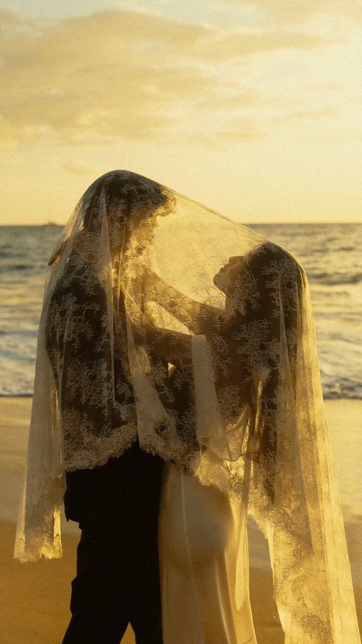 Wedding couple under a lace veil on a beach during golden hour sunset.
