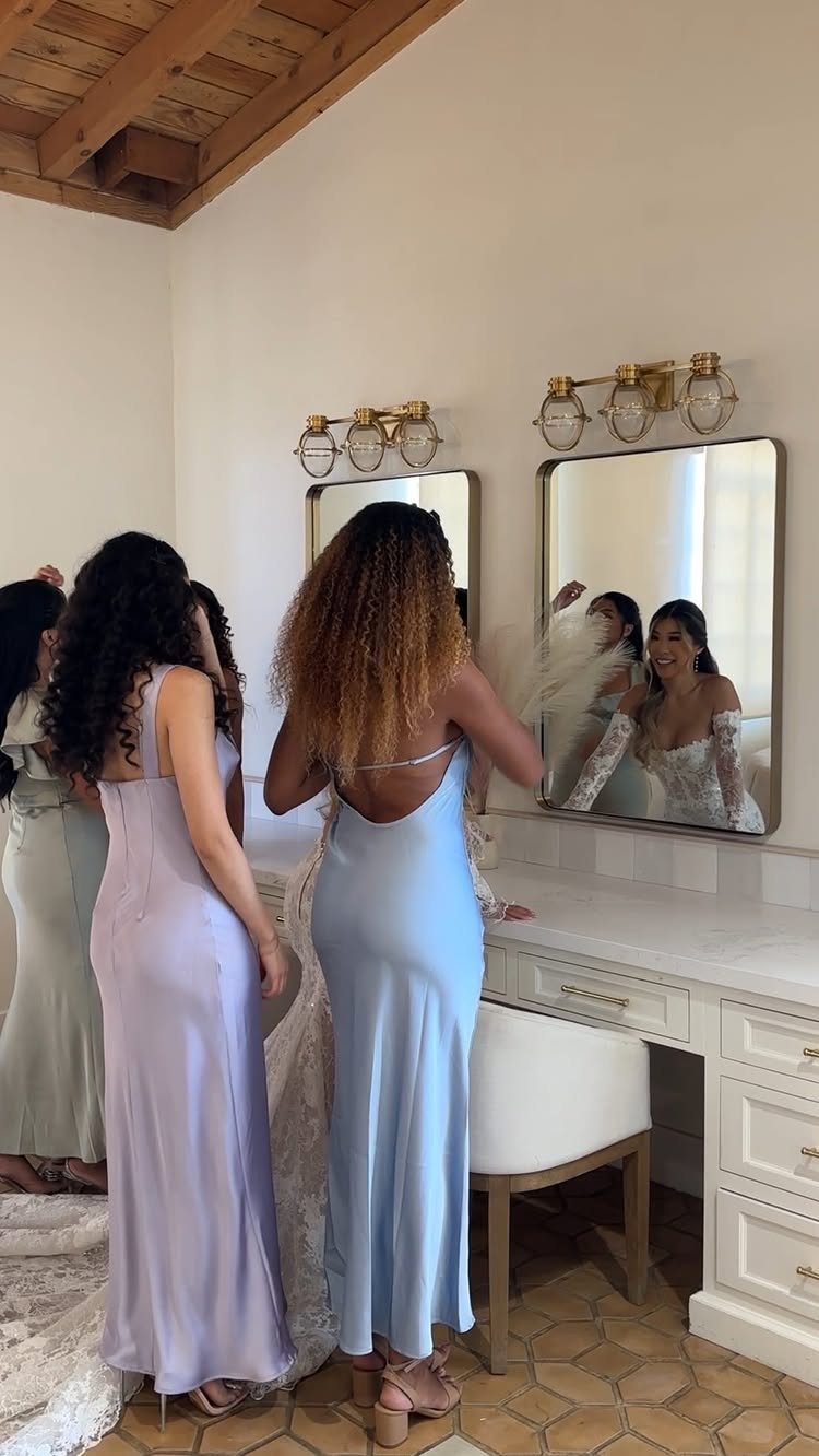 Bridesmaids in satin dresses standing by a white vanity mirror getting ready.