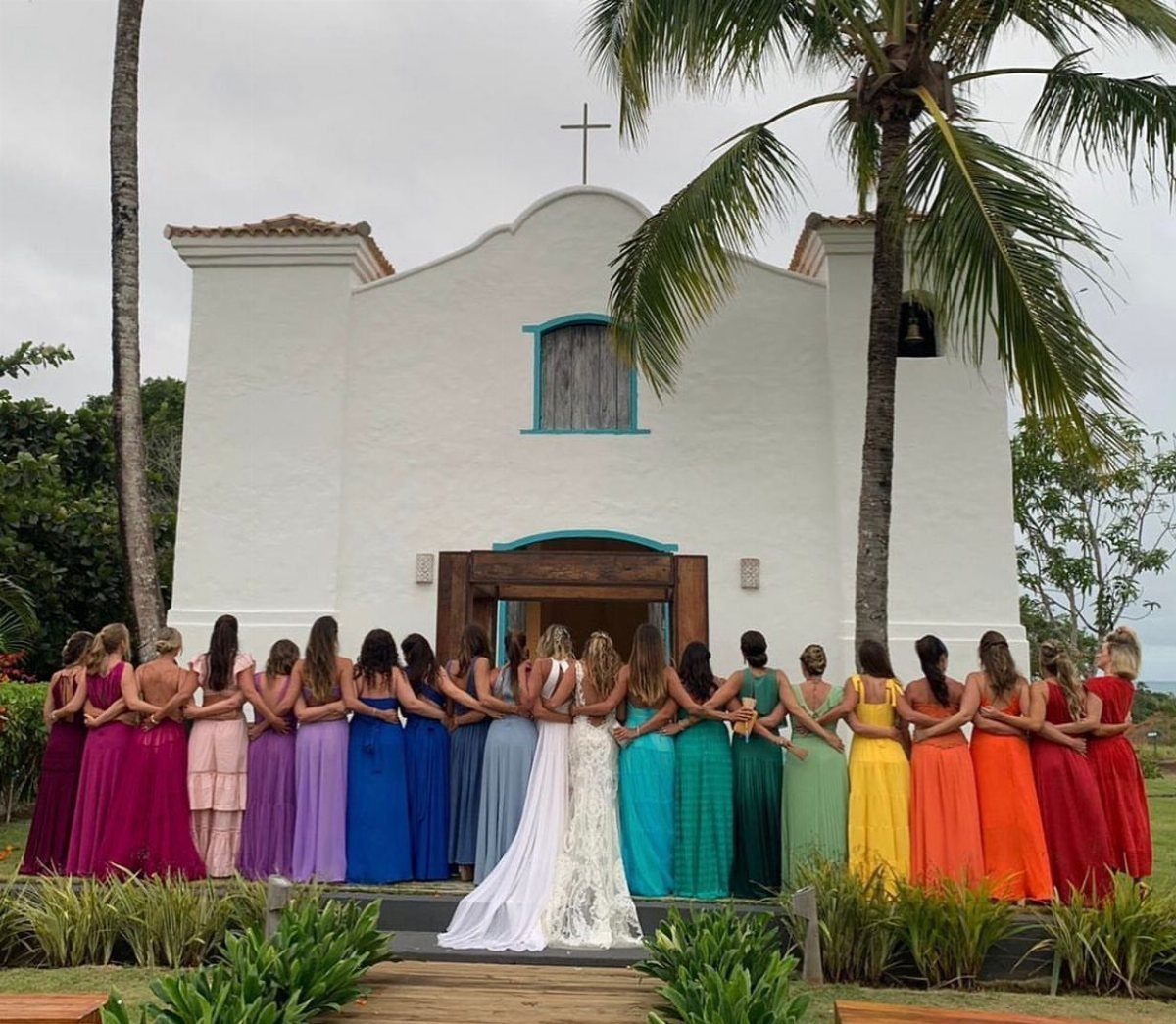 9 Uncommon Wedding Colors for Couples Who Skip the Usual 3 Bridesmaids in rainbow colored dresses standing before a white church with palm trees.