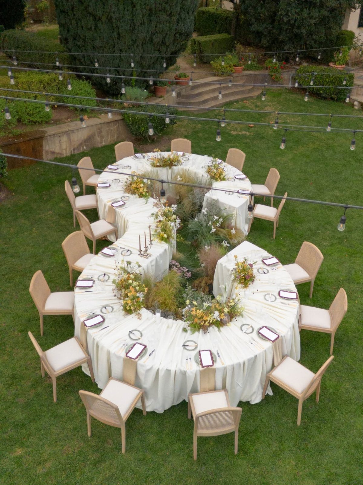 S-shaped white head table with floral arrangements and beige chairs on a green lawn