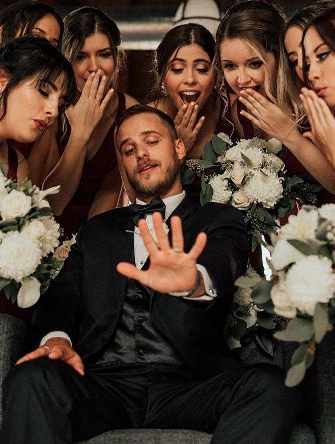 Groom in black tuxedo surrounded by five bridesmaids in red dresses with white bouquets