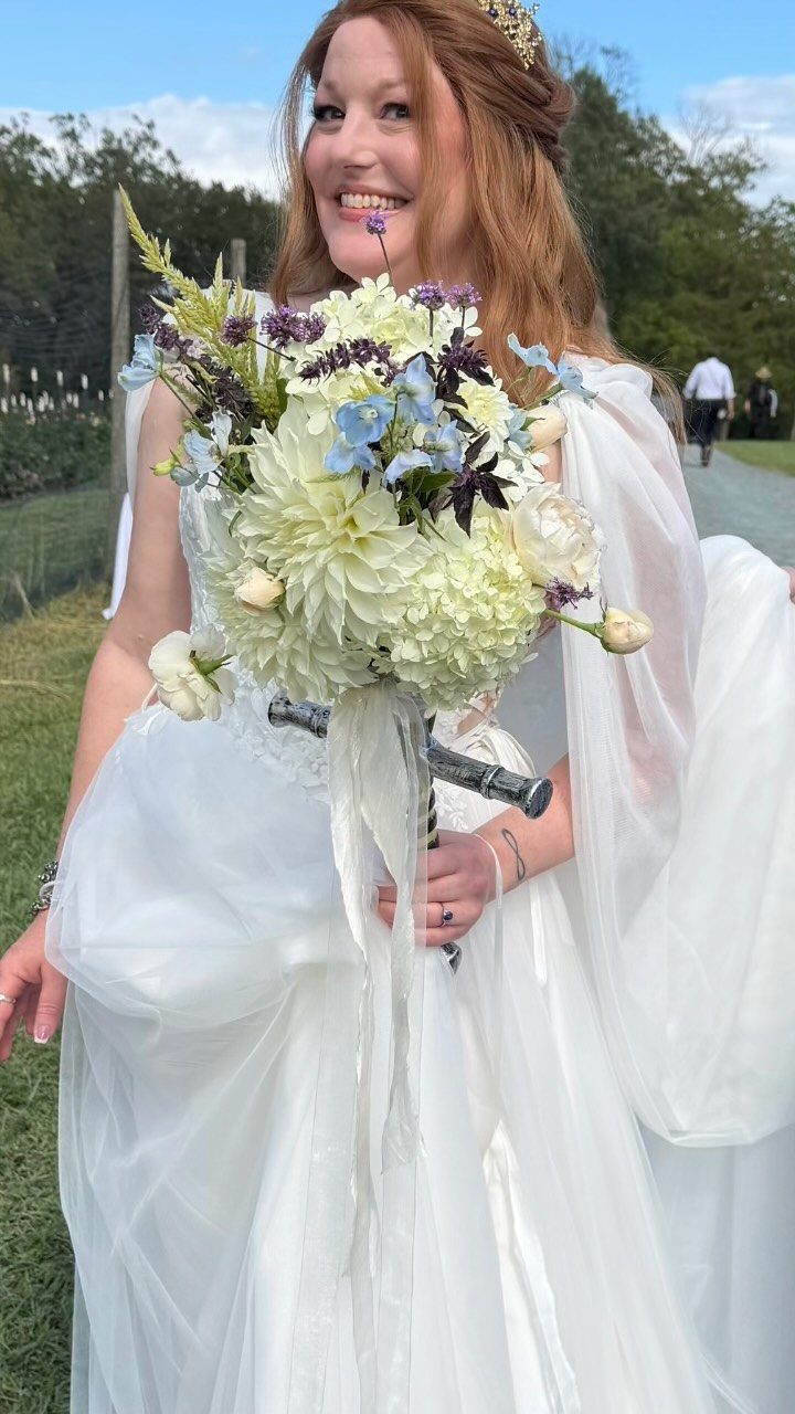Bride with red hair and gold crown holding a large floral bouquet outdoors.