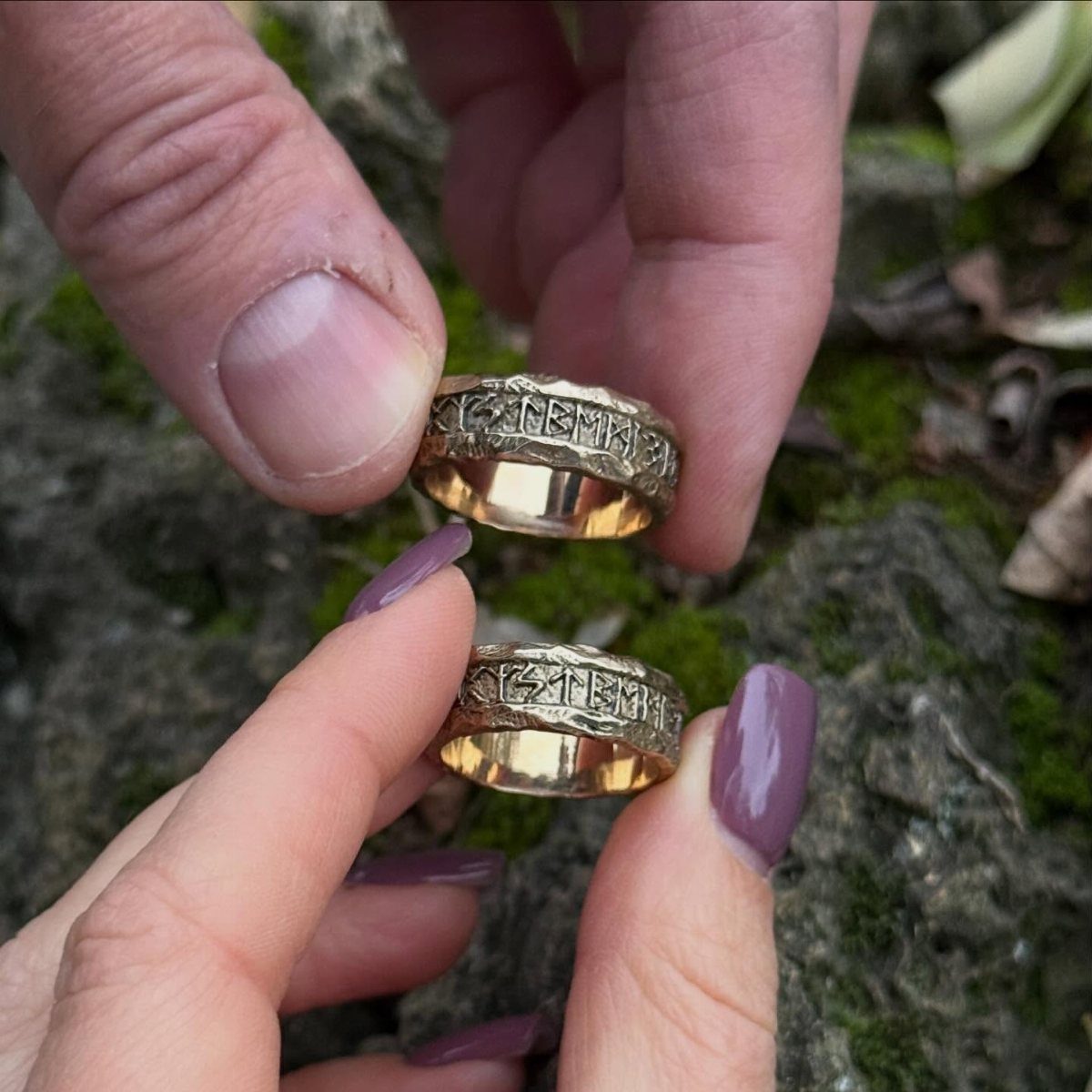 Two gold wedding rings with engraved Norse runes held by two people outdoors.