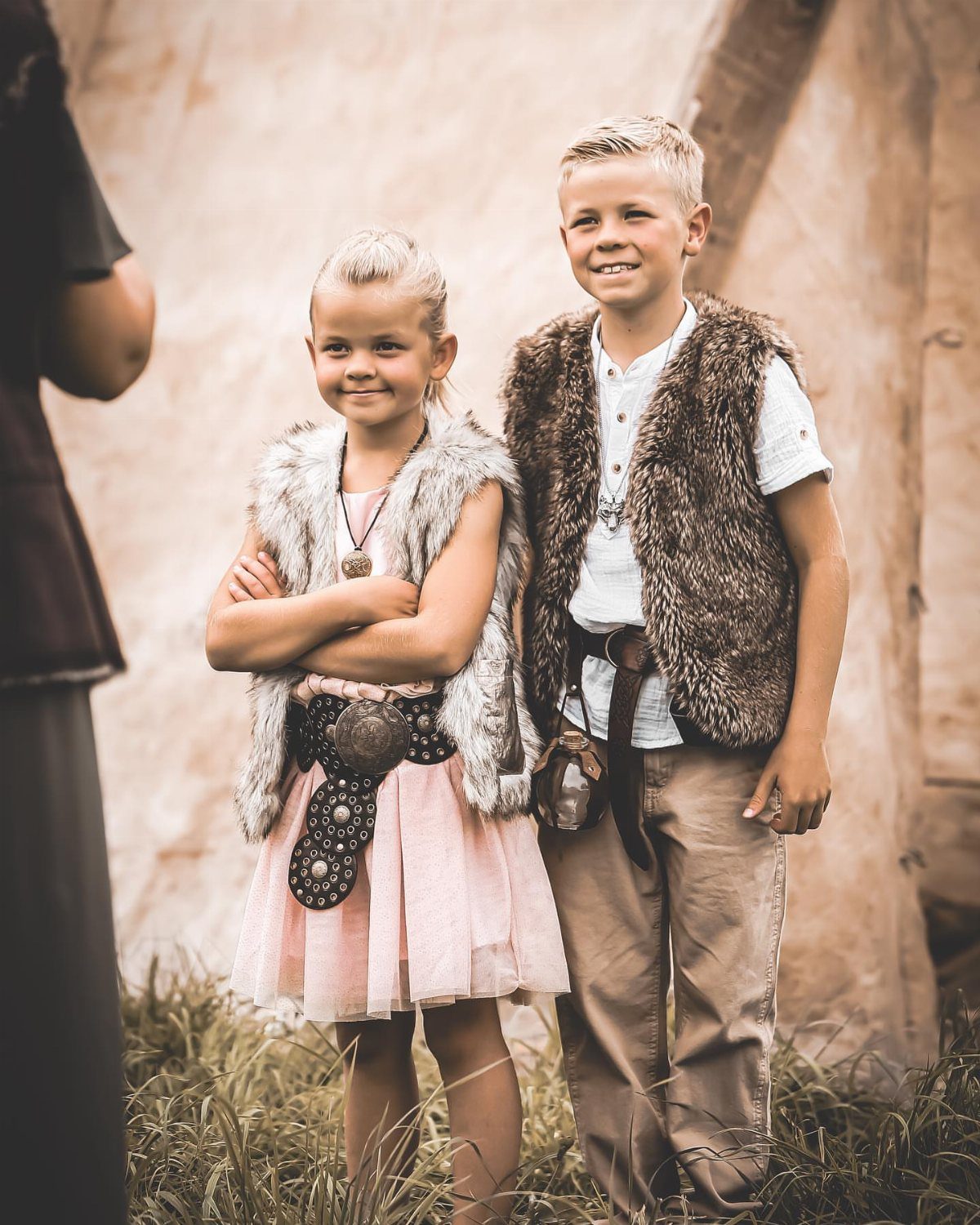Two children wearing Viking-style faux-fur vests and traditional accessories standing outdoors.