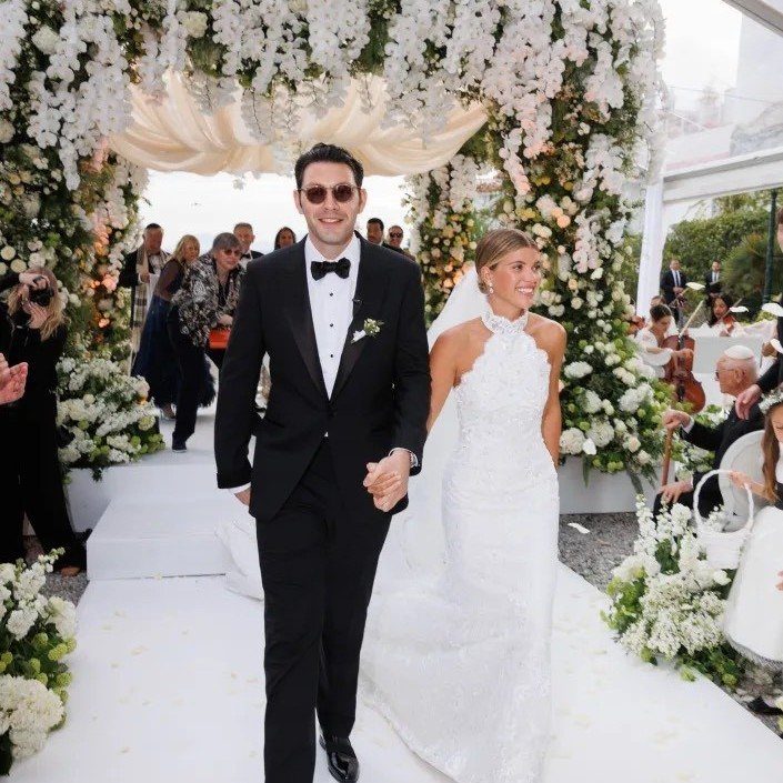 Vintage Chanel Wedding Dress Ideas: 8 Bridal Looks 6 Bride in white lace dress and groom in tuxedo walking under floral arch.