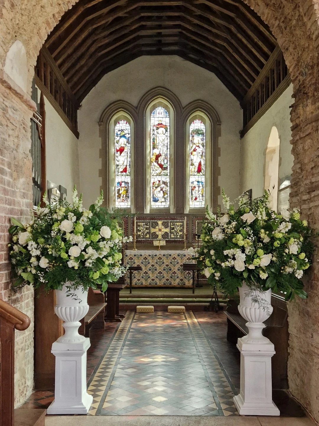5 Wedding Arch Without Flowers Ideas for Your Ceremony 3 White floral arrangements in decorative urns on pedestals inside a church aisle.
