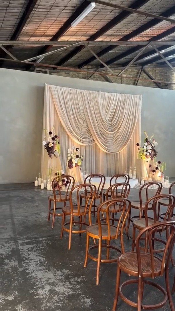 19 Wedding Backdrop Decorations for Any Ceremony Style 4 Cream draped fabric backdrop with floral arrangements and wooden chairs for a wedding ceremony.