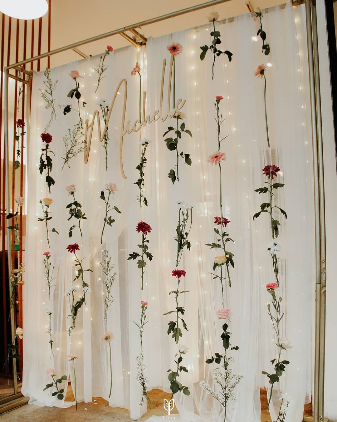 19 Wedding Backdrop Decorations for Any Ceremony Style 5 White fabric backdrop with hanging floral stems, fairy lights, and a gold name sign.