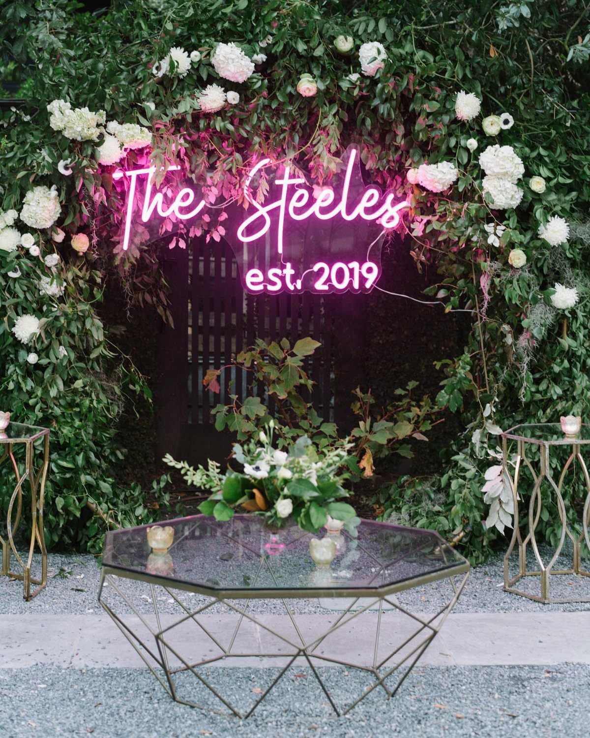 19 Wedding Backdrop Decorations for Any Ceremony Style 3 Pink neon sign with wedding names set against a floral greenery backdrop wall
