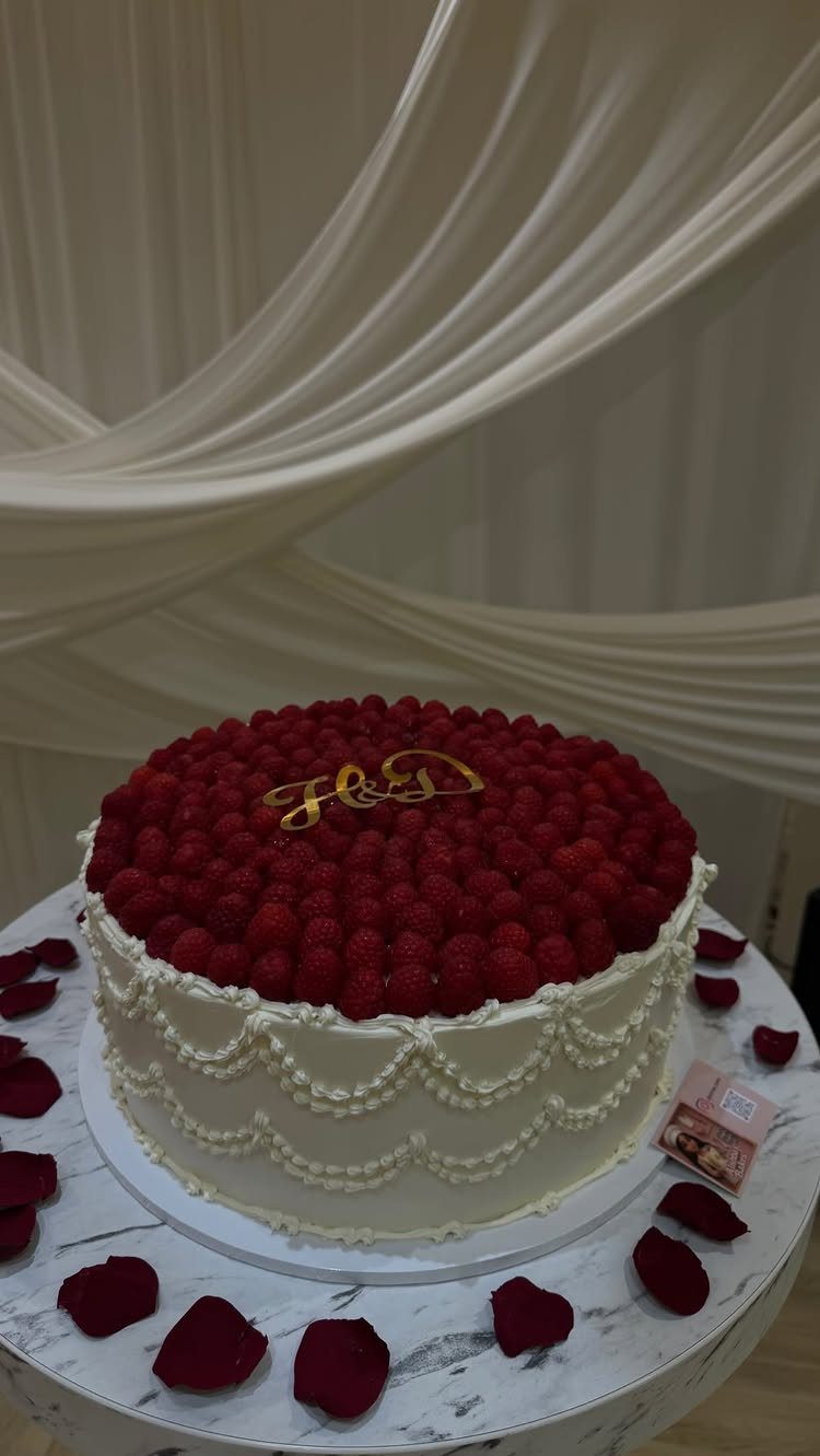 Round white wedding cake topped with raspberries and gold initials on marble table