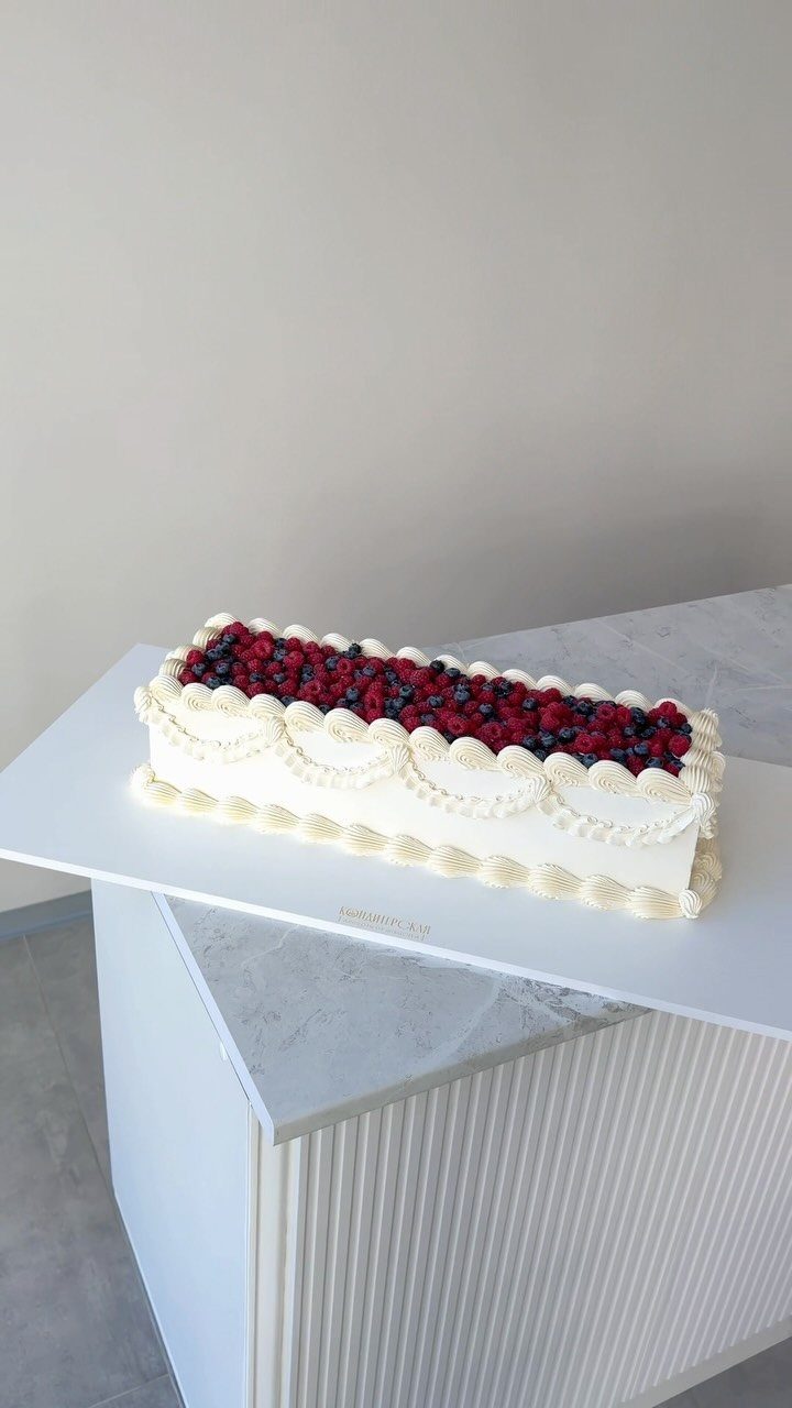 Rectangular white frosted cake topped with fresh raspberries and blueberries on white platform