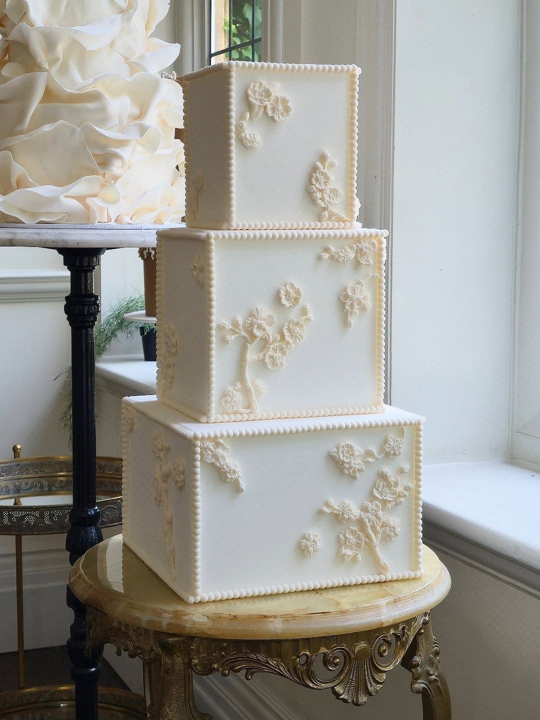 Three-tier white square wedding cake with floral frosting on an ornate gold table.